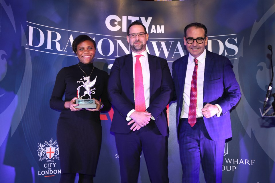 Feyi Osinuga accepts the award on behalf of Janus Henderson's Sarah de Lagarde, alongside Nick Carty of Eastern City BID and awards host Oli Barrett. City A.M. Dragon Awards 2024 at Mansion House. (Credit: Gretel Ensignia)